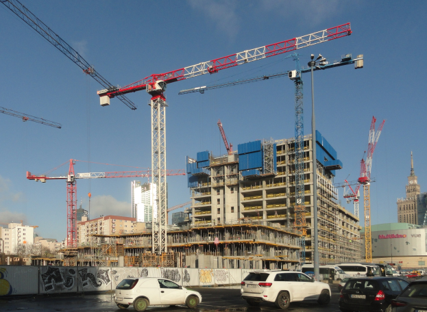 Baustelle mit Kränen, Fahrzeugen, Graffiti-beschmierten Wand, Gebäuden, Bäumen und bewölktem Himmel.