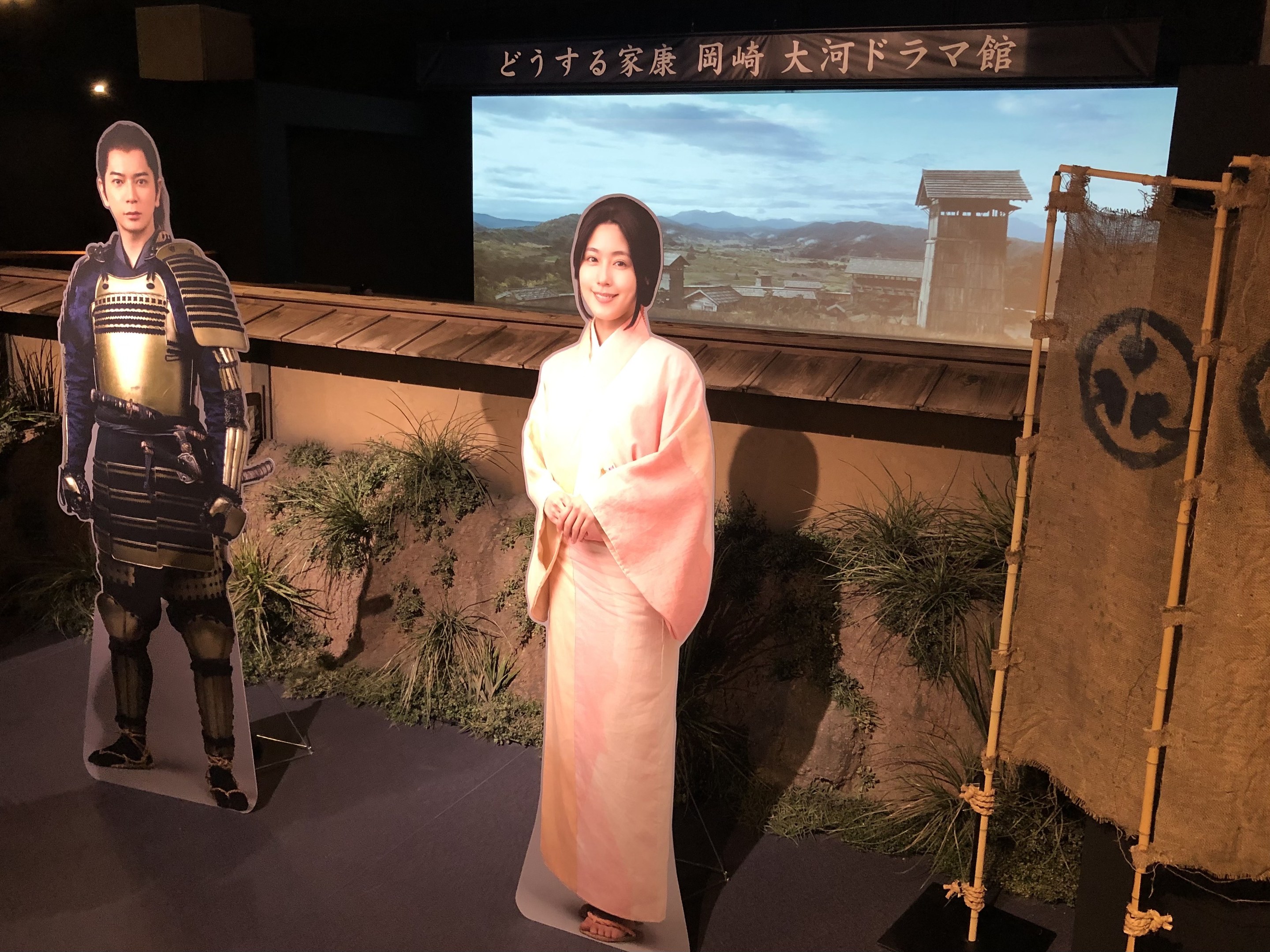Eine Frau in einem rosafarbenen Kimono und ein Mann in einem Samurai-Kostüm, die gemeinsam in einer Szene eines japanischen Films lächeln, mit Pflanzen, einem Bildschirm und Lichtern im Hintergrund.