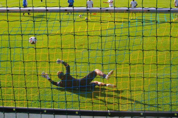 Ein Fussballtorhüter beim Abwehren eines Schusses vor einem Tor, mit einem Netz im Vordergrund und einigen Menschen im Hintergrund, während mehrere Fussbälle auf dem Boden liegen.