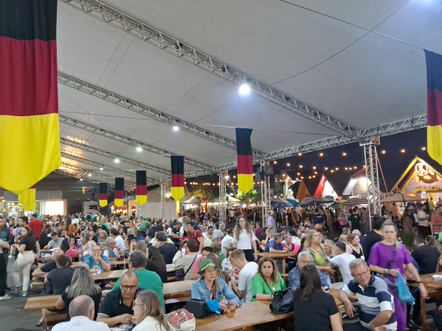 Eine große Gruppe von Menschen sitzt an Tischen unter einem Zelt auf dem Oktoberfest in München, Deutschland, während einige stehen und Taschen auf den Tischen liegen, während Zelte, Lichter und Fahnen eine festliche Atmosphäre schaffen.