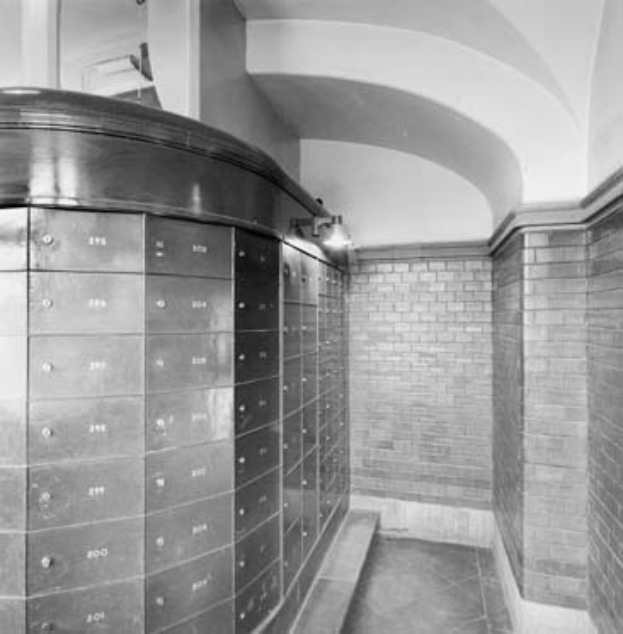 Ein Schwarz-Weiß-Foto eines Banktresorraums mit einer Wand voller Schließfächer, die mit Münzen und anderen Gegenständen gefüllt sind, einem Boden, einer Säule und einer Decke.