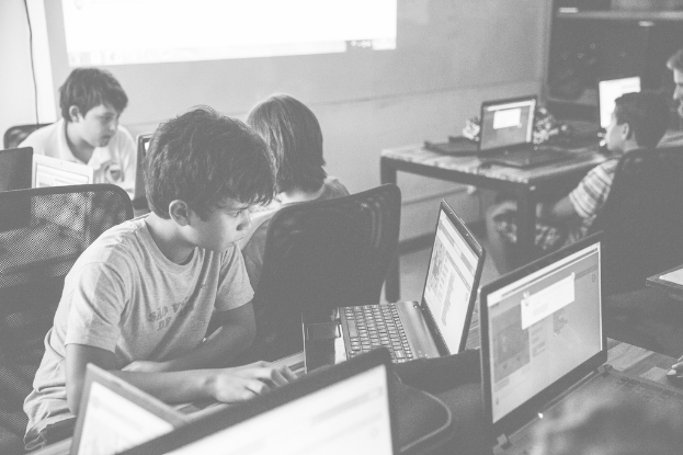 Ein Tisch mit schwarzen Laptops und schwarzen Stühlen, darauf sitzende Kinder, vor einer weißen Wand mit weißer Projektion im Hintergrund.