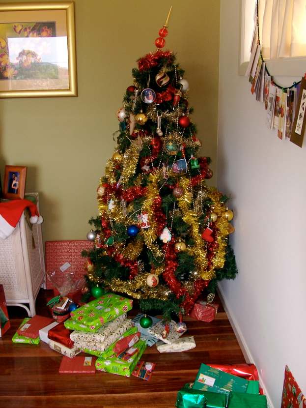 Ein geschmückter Weihnachtsbaum mit Geschenken am Fuße, ein Foto Rahmen an der linken Wand und ein Vorhang am rechten Fenster.