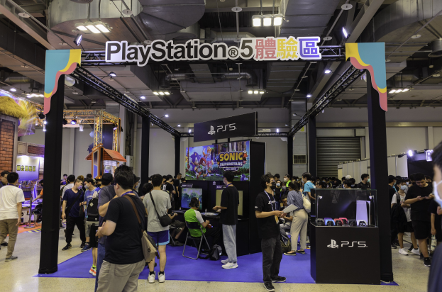 Eine Gruppe von Menschen an einem PlayStation-Stand während der Tokyo Games Expo, wobei einige Taschen tragen oder auf Stühlen sitzen, umgeben von Tafeln, Deckenleuchten, einem Bildschirm und einem Tisch mit Gegenständen auf der rechten Seite.