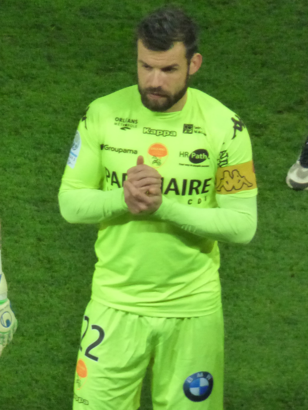 Ein Mann in einem gelben Torwarttrikot, einem grünen T-Shirt und Shorts steht auf einem Fußballfeld und klatscht in die Hände, wobei auf der linken Seite des Bildes das Bein einer anderen Person zu sehen ist.