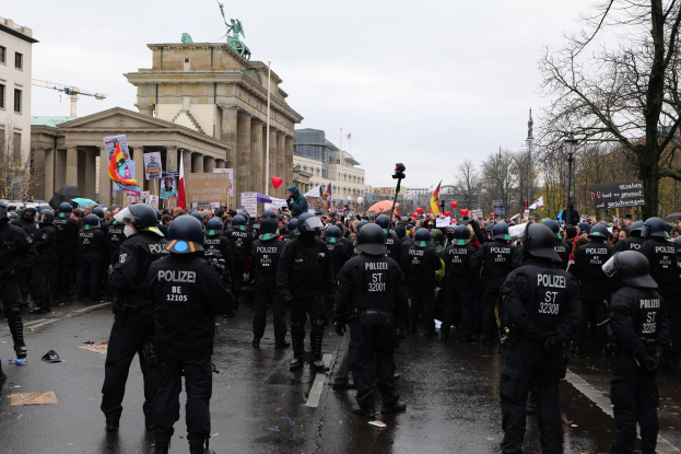 Eine große Gruppe von Polizisten steht vor einer Menge mit Schildern, Ballons und Helmen, im Hintergrund sind Gebäude, Bäume, eine Statue, ein Kran und ein bewölkter Himmel zu sehen.