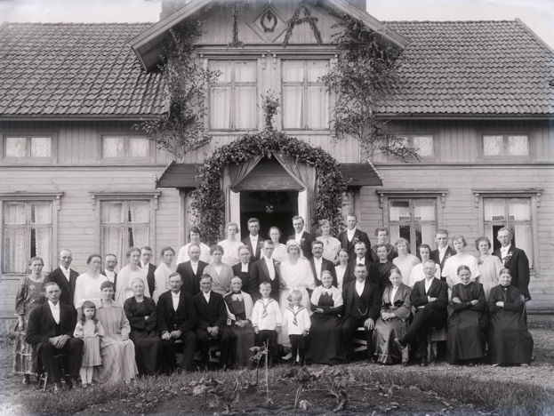 Schwarzes und weißes Foto einer großen Gruppe von Menschen, die vor einem Haus mit reichlicher Vegetation und einem sichtbaren Himmel posieren.