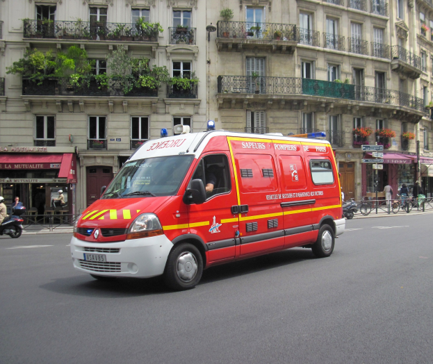 Rettungswagen steht auf der Straße vor einem Gebäude mit Fenstern, Geländern, Pflanzen und anderen Gegenständen, mit einigen Menschen und Fahrrädern im Hintergrund.