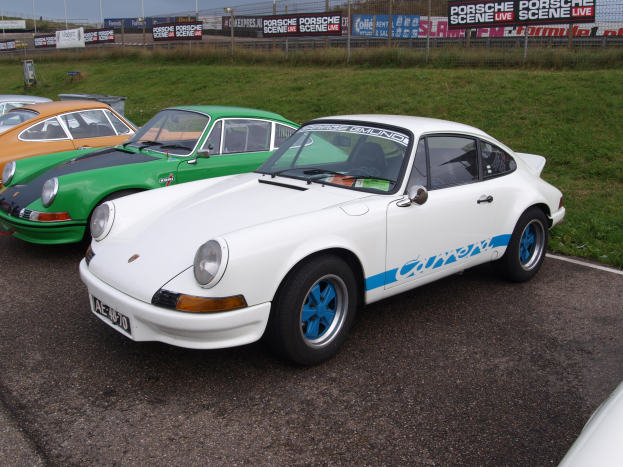 Drei Porsche 911s nebeneinander auf einem Parkplatz mit Gras, einem Zaun, Bannern, Laternenmasten und einem bewölkten Himmel im Hintergrund geparkt.