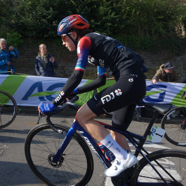 Eine professionelle Radrennfahrerin, Katie Archibald, fährt mit Helm, Schutzbrille, Handschuhen und Schuhen ein Fahrrad auf einer Straße neben einer Menschenmenge. Im Hintergrund gibt es ein Banner, eine Wand, Bäume und ein Gebäude.