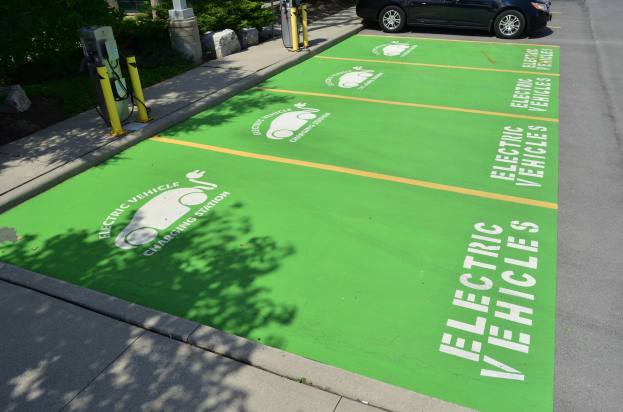 Elektroauto an einer Ladestation mit grünem Bodenmatte mit Text und Bildern in einer Parkanlage mit Bäumen, Pflanzen und Masten.