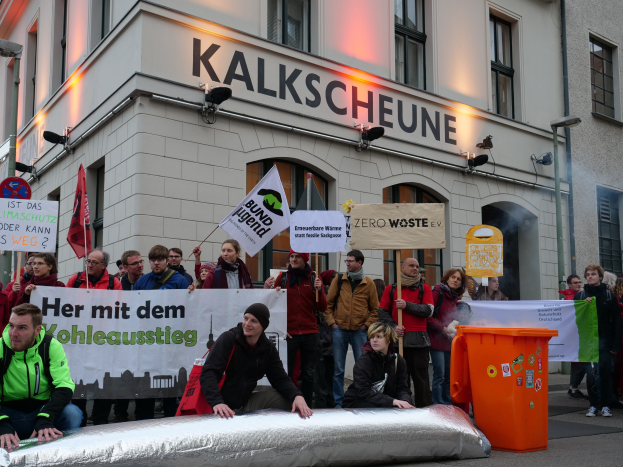 Eine Gruppe von Menschen mit Protestschildern und Plakaten vor einem Gebäude, mit zwei Personen im Vordergrund auf einem Gegenstand sitzend und einem Müllcontainer rechts daneben, vor einem Hintergrund von Gebäuden mit Fenstern und Schildern in Deutschland.