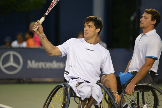 Zwei Männer im Rollstuhl, einer hält einen Stock, beide tragen weiße T-Shirts, vor einer Wand mit dem Mercedes-Logo und Text, mit Menschen im Hintergrund.