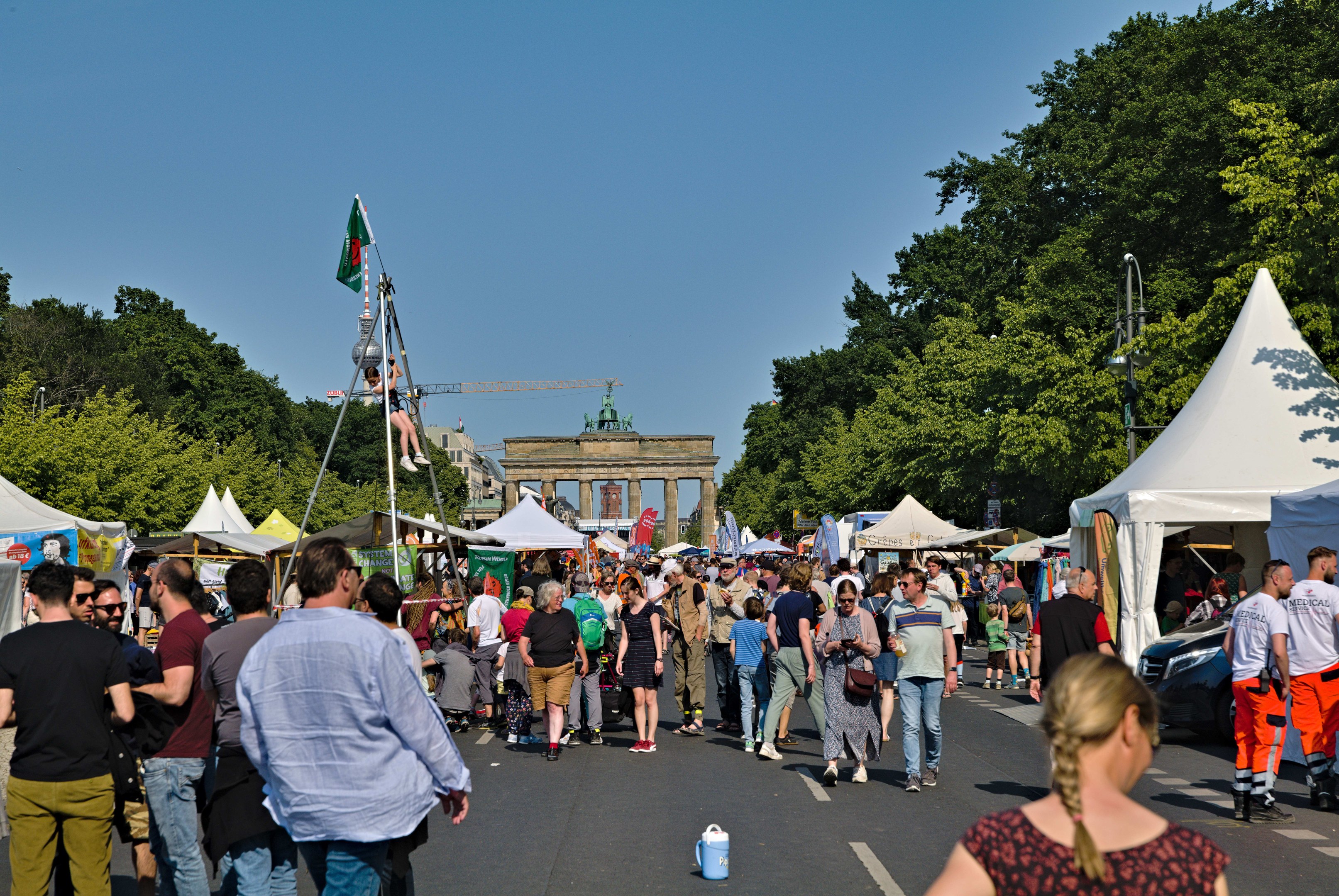 Eine Menschenmenge geht eine Straße mit Zelten, Fahrzeugen und Bäumen entlang, mit einem Bogen und einem klaren blauen Himmel im Hintergrund und Flaggenmasten auf der linken Seite, die auf ein Oktoberfest hinweisen.