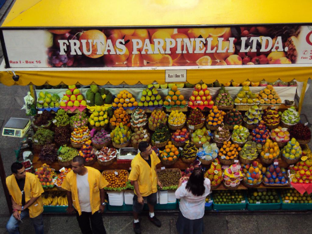 Ein Marktstand mit Körben voller Früchte, einem Banner und einer Tafel oben sowie einer Waage links und vier Personen unten auf dem Weg.
