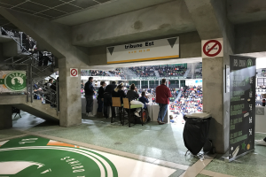 Eine Gruppe von Menschen steht vor einem sitzenden Publikum in einem Stadion, mit Schildern, die "Boston Celtics vs New York Jets", ein Müllcontainer, eine Texttafel, eine Treppe mit Geländern und eine Decke mit Deckenleuchten anzeigen.
