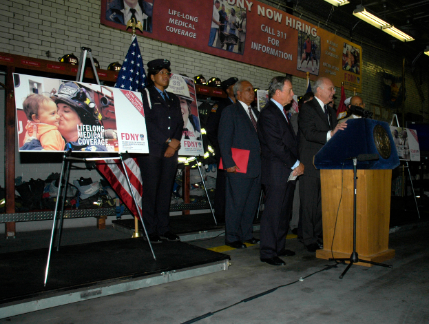 Eine Gruppe von Menschen, die in der Nähe eines Podiums mit Mikrofonen, Schildern auf Ständern, Fahnen und Schildern an der Wand stehen, alles unter hellen Lichtern.
