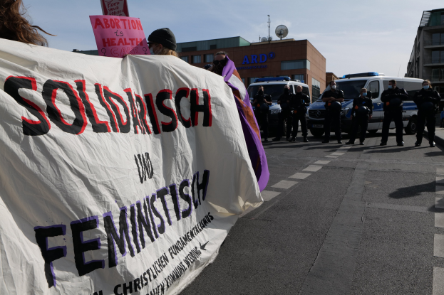 Gruppe von Menschen marschiert auf einer Straße mit einem Banner "Solidarität und Feminismus", mit parkenden Fahrzeugen, Gebäuden, einer Schüsselantenne und einem klaren blauen Himmel im Hintergrund.