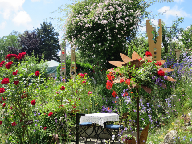 Ein Garten auf der Chelsea Flower Show mit bunten Blumen und Pflanzen, einem Tisch mit einer Decke, Stühlen, Bäumen und einem bewölkten Himmel im Hintergrund.