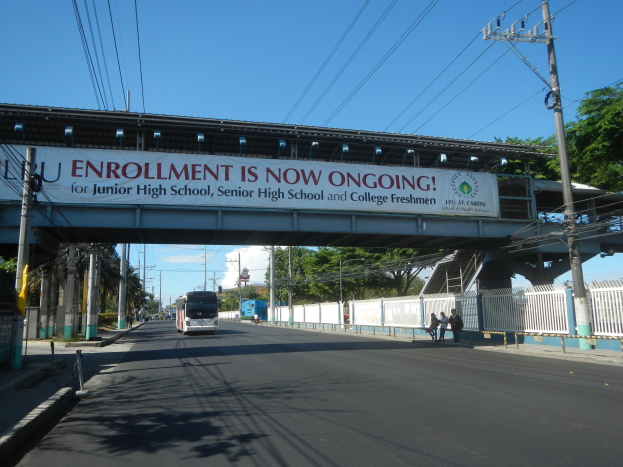 Ein Bus fährt unter einer Brücke mit einem Schild "Anmeldung läuft" vorbei, Menschen gehen auf dem Gehweg darunter, Strommasten mit Drähten, Bäume und ein klarer blauer Himmel im Hintergrund.