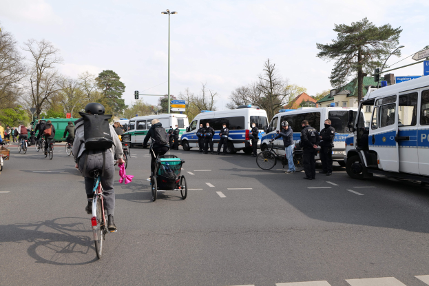 Eine Gruppe von Menschen, die auf Fahrrädern eine Straße entlangfahren, gesäumt von Polizeiwagen und anderen Fahrzeugen, einige tragen Helme und haben Taschen oder schieben Kinderwagen.