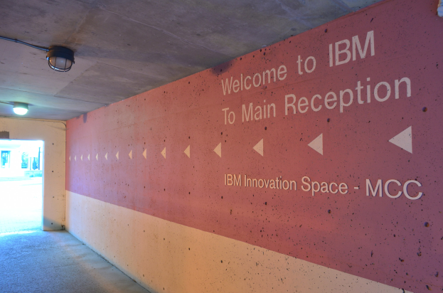 Eine Wand mit dem Text "Willkommen bei IBM" beleuchtet von Deckenlampen, mit einer Tür im Hintergrund beschriftet "IBM Innovation Space MCC."