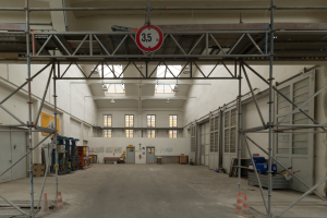 Großer Lagerraum mit ausgedehnten Gerüsten, Verkehrskegeln, Metallstangen, einem Schild, Maschinen, einer Tür, Fenstern, Wänden, Deckenlampen und einem Dach.