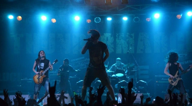 Eine Gruppe von Musikern, die auf einer hell erleuchteten Bühne spielen, wobei einige Gitarren spielen, einer ein Mikrofon hält und ein anderer im Hintergrund Schlagzeug spielt, während die Hände des Publikums unten zu sehen sind.