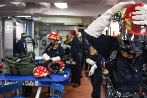 Eine Gruppe von Menschen in Helmen und Handschuhen steht um einen Tisch mit Helmen und Taschen, Stühle sind im Raum verteilt, mit Türen und Deckenlampen im Hintergrund.