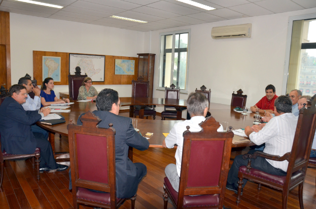 Eine Gruppe von Menschen sitzt an einem Tisch mit Papieren und Gläsern, im Hintergrund sind eine Klimaanlage, eine Anschlagtafel und Karten an der Wand zu sehen.