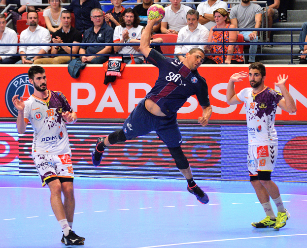 Männer beim Handballspielen auf einem Court mit einem Ball in der Mitte, Zuschauern im Hintergrund und verstreuten Kleidungsstücken auf dem Court, mit einem Banner, auf dem "Futsal-Weltmeisterschaft 2015 - Paris Saint-Germain vs. Olympique Lyon" steht.