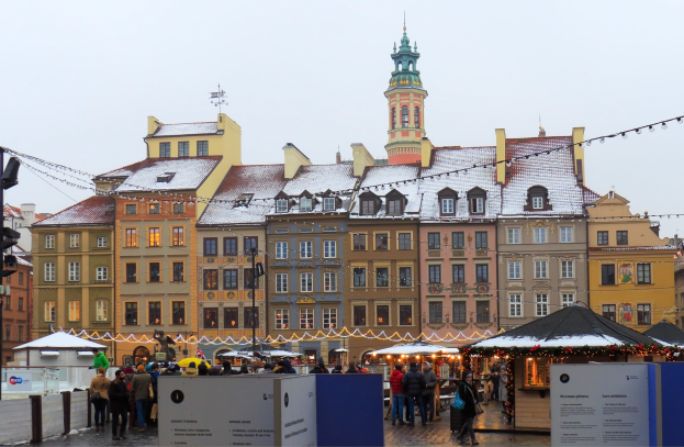 Ein lebendiger Weihnachtsmarkt im alten Stadtzentrum von Warschau mit Menschen um beleuchtete und dekorierte Stände, Gebäuden mit Fenstern im Hintergrund und einem Verkehrslichtmast auf der linken Seite.