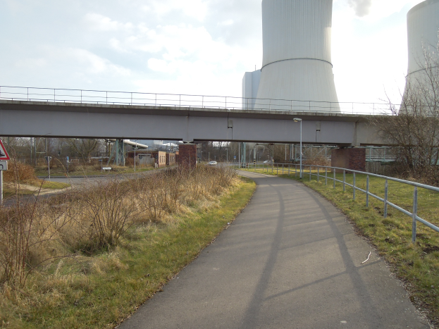 Eine von Gras und Pflanzen gesäumte Straße führt zu einer Brücke mit Geländern, Laternenmästen und einem Schild, im Hintergrund eine Atomkraftwerk mit Rauch aus dem Schornstein unter einem bewölktem Himmel.