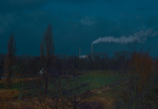 Outdoorszene mit einem bewölkten Himmel, einer rauchenden Fabrik, ein paar Häusern und einem grasbewachsenen Bereich, der mit Bäumen und Pflanzen gefüllt ist.