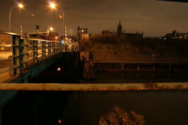 Eine Brücke spannt einen Fluss mit zahlreichen Straßenlaternen darauf und Gebäude sind im Hintergrund sichtbar.