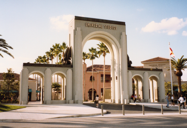Eingang zu Universal Studios Hollywood mit einem Bogen mit Text, einer Statue, Menschen, Bäumen, Gebäuden, einem Fahnenmast mit einer Flagge und einem bewölkten Himmel.
