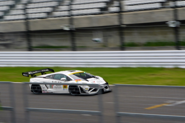 Weißer Sportwagen auf einer Rennstrecke umgeben von einem Zaun und Gras, mit einem unscharfen Hintergrund, der hohe Geschwindigkeit andeutet.