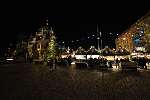 Ein nächtlicher Weihnachtsmarkt in Berlin, Deutschland, mit Menschen zwischen Gebäuden, Straßenlaternen, geschmückten Bäumen und verschiedenen Gegenständen auf dem Boden unter einem bewölkten Himmel.