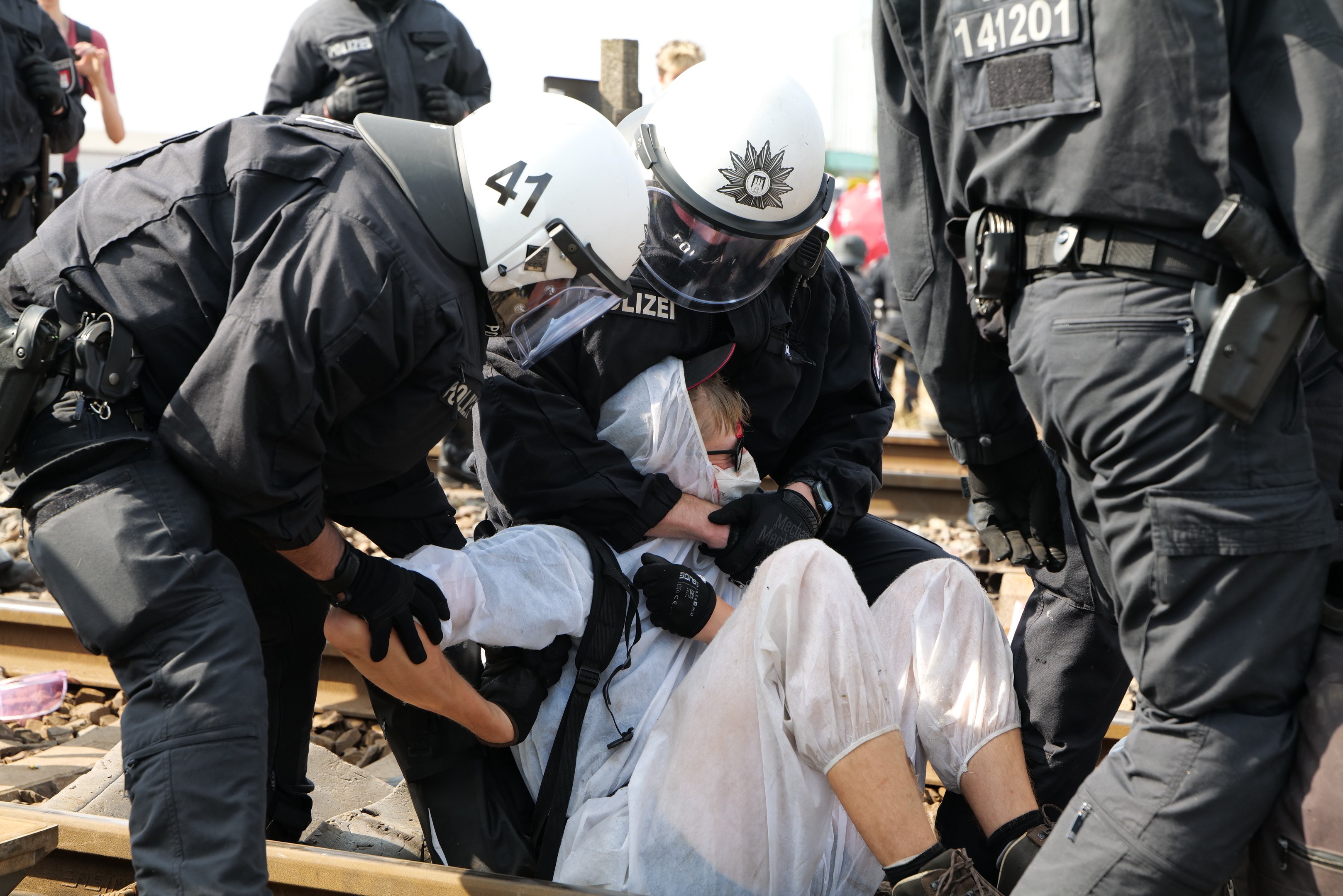 Polizeibeamte in Einsatzausrüstung verhaften einen Mann in einem weißen Kleid auf einem Bahngleis, mit anderen im Hintergrund.