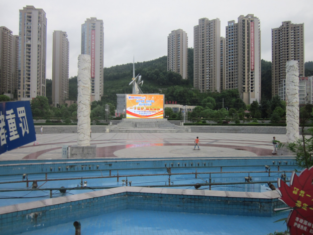 Großer Swimmingpool umgeben von hohen Gebäuden in einer städtischen Umgebung, mit ein paar Menschen in der Nähe, einer Fahne auf der linken Seite, Bäumen im Hintergrund und einem klaren blauen Himmel.