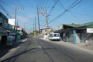 Ein Bus fährt auf einer Straße mit Strommasten und Kabeln entlang, flankiert von Gebäuden, mit Passanten auf den Gehwegen und Bäumen im Hintergrund unter einem klaren blauen Himmel.
