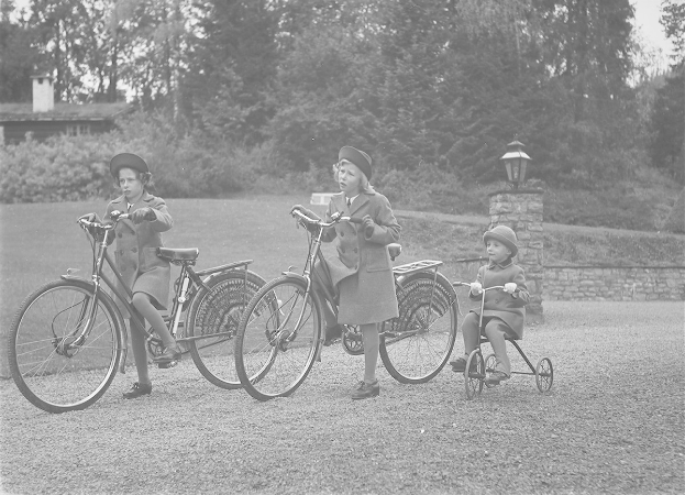 Drei Kinder in Mänteln und Mützen fahren Fahrräder und ein Dreirad in einem Park mit Bäumen, Pflanzen, einem Haus und einem Laternenpfahl in Schwarz-Weiß.