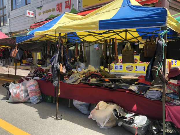 Ein Straßenmarkt mit Zelten, die verschiedene Gegenstände wie Taschen und Hüte präsentieren, vor einem Hintergrund aus Gebäuden mit Fenstern und Texttafeln.
