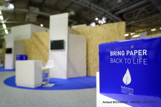 Ein Messestand mit einem blauen und weißen Schild "Bring Paper Back to Life" auf der rechten Seite, im Hintergrund sind Stühle, Tische, ein Fernseher und Deckenleuchten zu sehen.
