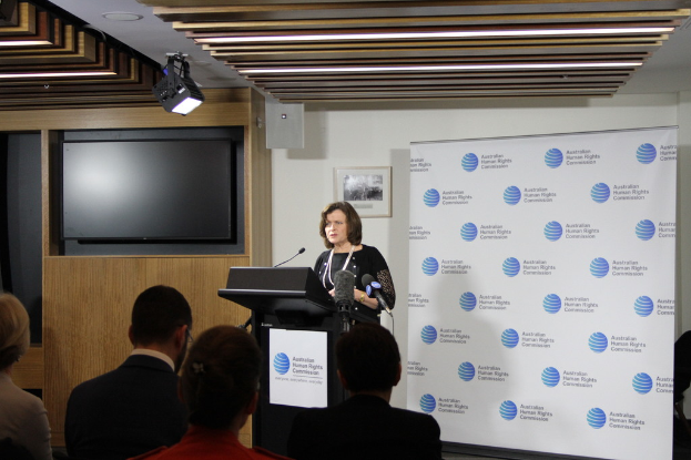 Eine Frau steht auf einer Bühne und spricht in ein Mikrofon, vor ihr sitzen Zuschauer, hinter ihr eine Wand mit einem Banner und ein Fernseher an der Holzwand, an der Decke Lampen.