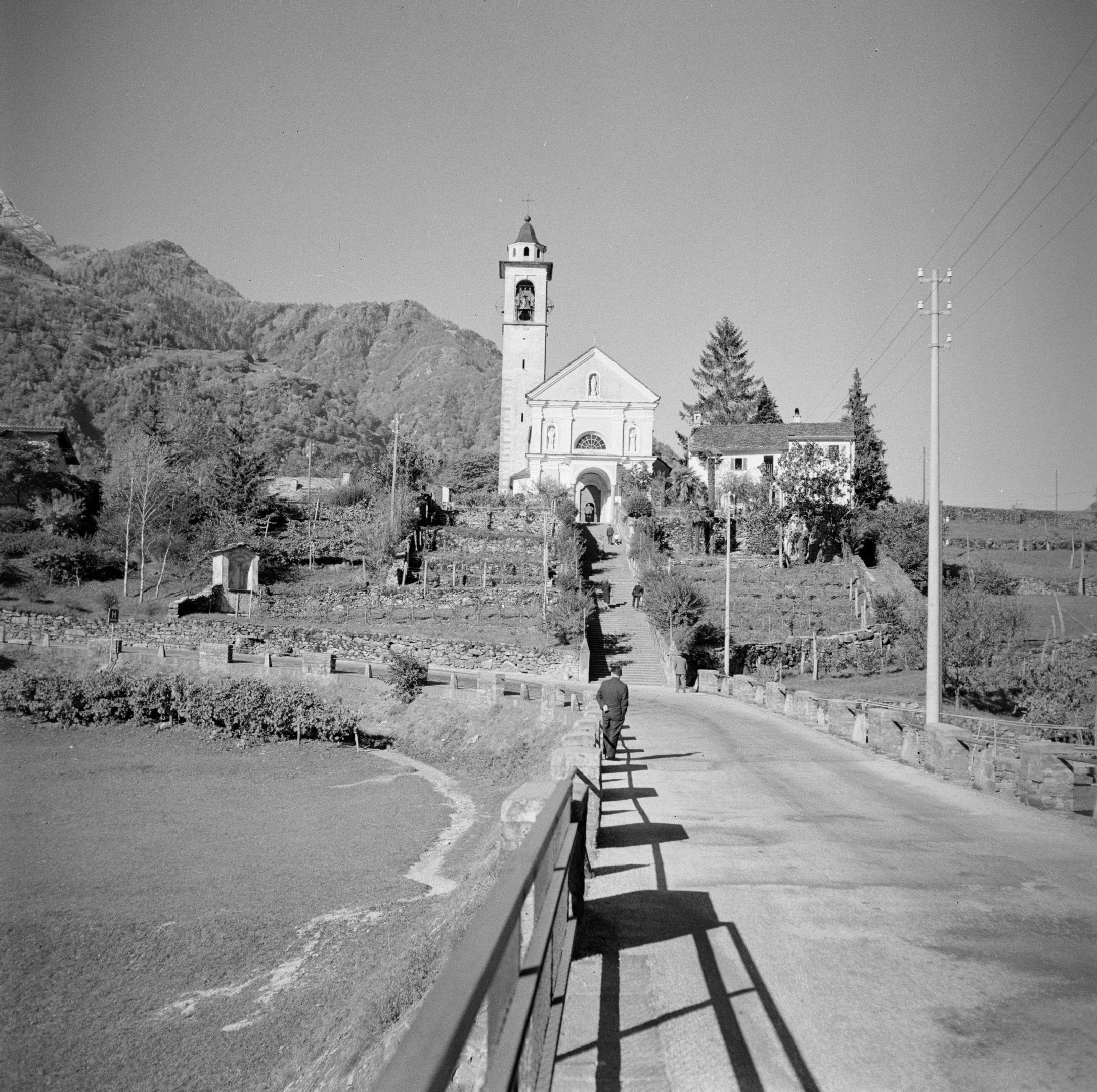 Schwarzes und weiße Foto einer Person, die ein Fahrrad auf einer Straße mit Geländern fährt, umgeben von Bäumen, Gebäuden, Masten, Drähten, Höhen und einem klaren Himmel.