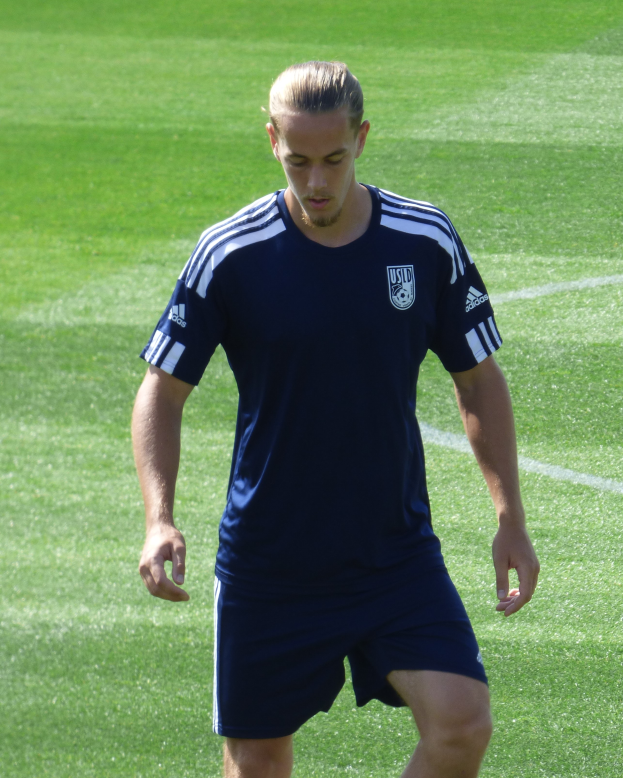 Ein Mann in einem blauen T-Shirt und Shorts läuft selbstbewusst über ein saftiges grünes Feld und sieht aus wie ein professioneller Fußballspieler mit entschlossener Miene.