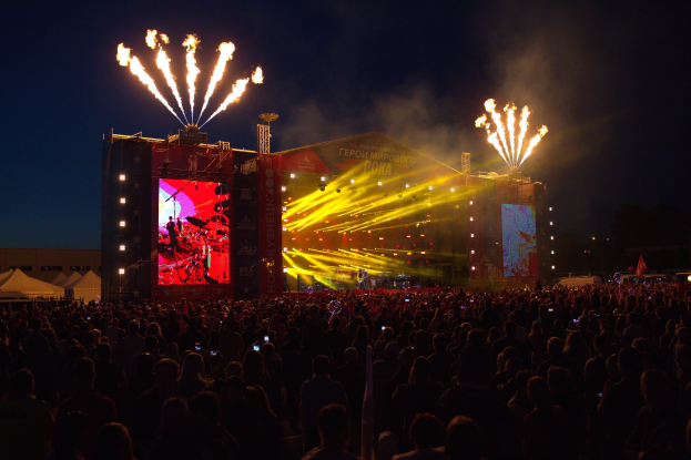 Großes Publikum bei Musikfestival mit beleuchteter Bühne und Performern, Bannern, Zelten links und Feuerwerk am dunklen Himmel.