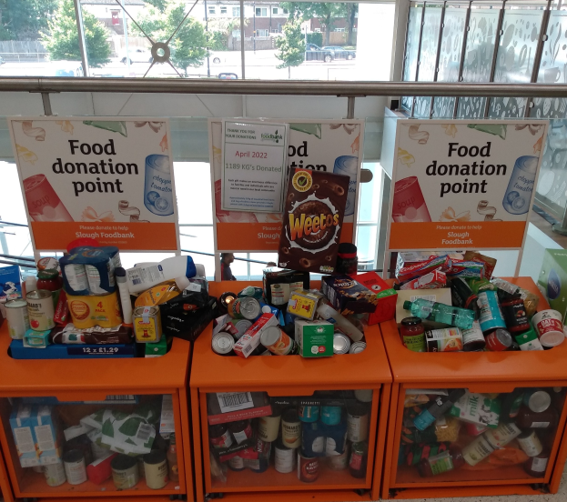 Orange Schränke mit Lebensmitteln wie Flaschen, Dosen und Kartons dienen als Spendenstation, daneben befinden sich Informationsschilder; draußen sind Fahrzeuge, Bäume, Gebäude und Himmel durch Glasfenster zu sehen.