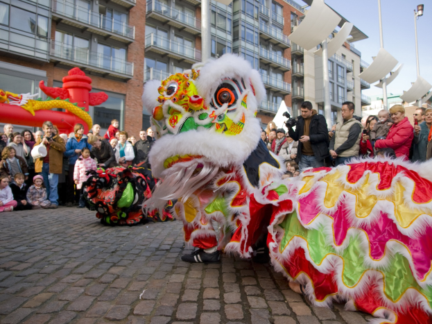 Ein lebendiges chinesisches Neujahrsfest in Amsterdam mit einer Löwen-Tanz-Show vor einer Zuschauermenge, darunter einige, die das Ereignis fotografieren, vor einer Kulisse aus Gebäuden, Laternenmasten und einem klaren blauen Himmel.